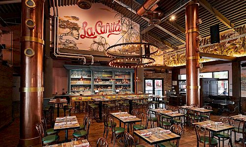 A cozy, industrial-style restaurant with rustic wooden tables, green chairs, exposed copper columns, warm lighting, and a mural-style bar area labeled &ldquo;La Cantina.&rdquo;