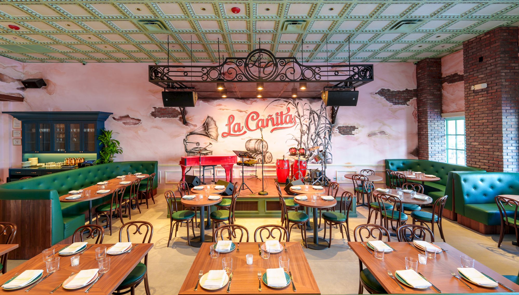 A cozy restaurant interior with brick walls, green booths, wooden tables set with plates and cutlery, and a stage with a mural reading &ldquo;La Cantina.&rdquo;