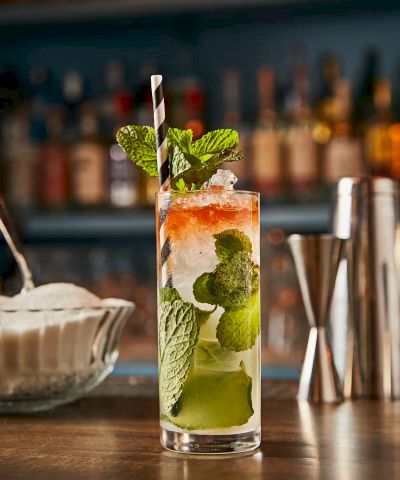 A tall mojito-style cocktail with mint, lime, and crushed ice, garnished with a mint sprig and a striped straw, set against a bar backdrop.