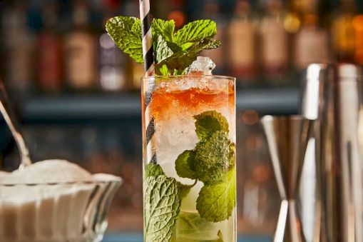 A tall mojito-style cocktail with mint, lime, and crushed ice, garnished with a mint sprig and a striped straw, set against a bar backdrop.