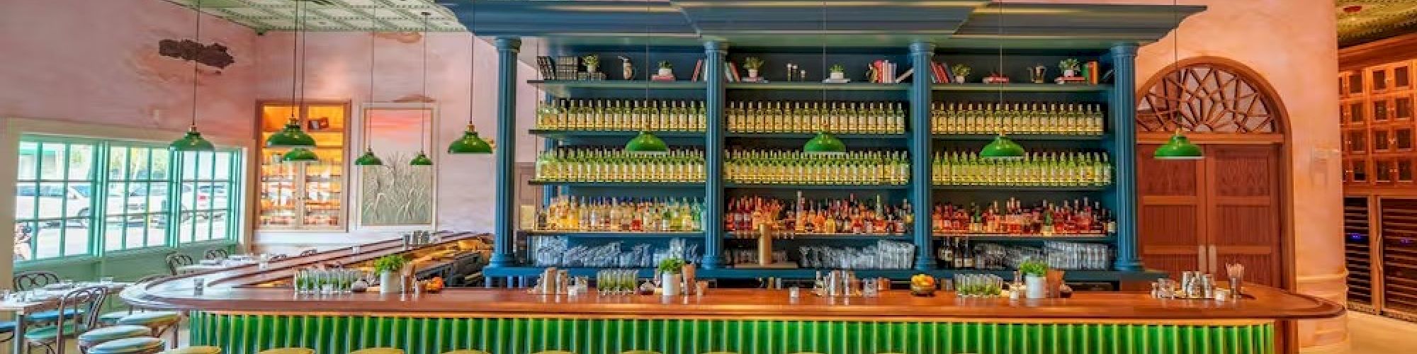 A stylish bar with a long green counter, blue shelves stocked with bottles, row of green-topped stools, warm lighting, and a vintage ceiling. End.
