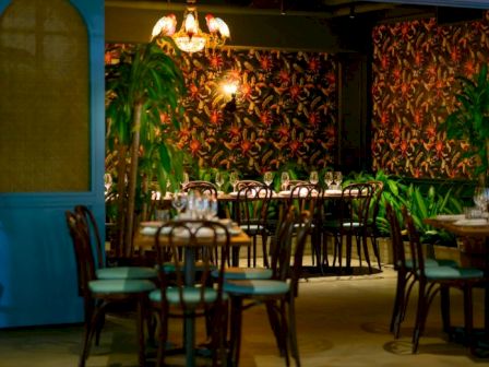 Cozy restaurant interior with palm-print wallpaper, teal chairs, wooden tables, warm lighting, and blue door at the left.