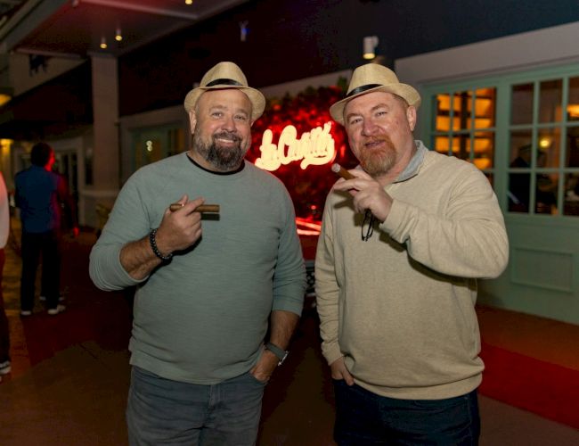 Two smiling men wearing matching beige hats pose with drinks at a lively bar, neon signage glowing behind them.
