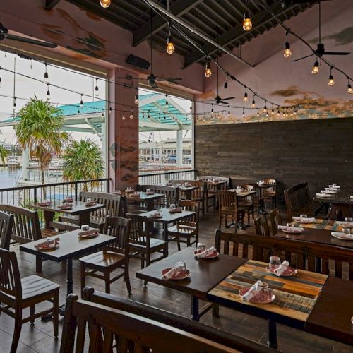 A stylish open-air restaurant with wooden tables, string lights, and a view of palm trees and water in the background, ready for dining.