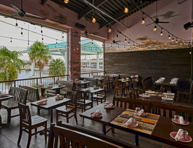 A stylish open-air restaurant with wooden tables, string lights, and a view of palm trees and water in the background, ready for dining.