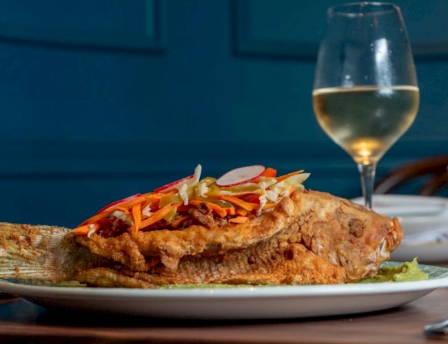 A large sandwich or open-faced dish topped with shredded vegetables on a plate, with a glass of white wine in the background.