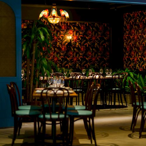 A stylish restaurant interior with dim lighting, patterned wallpaper, plants, and neatly set tables. Top it at 140 characters, always ending the sentence.