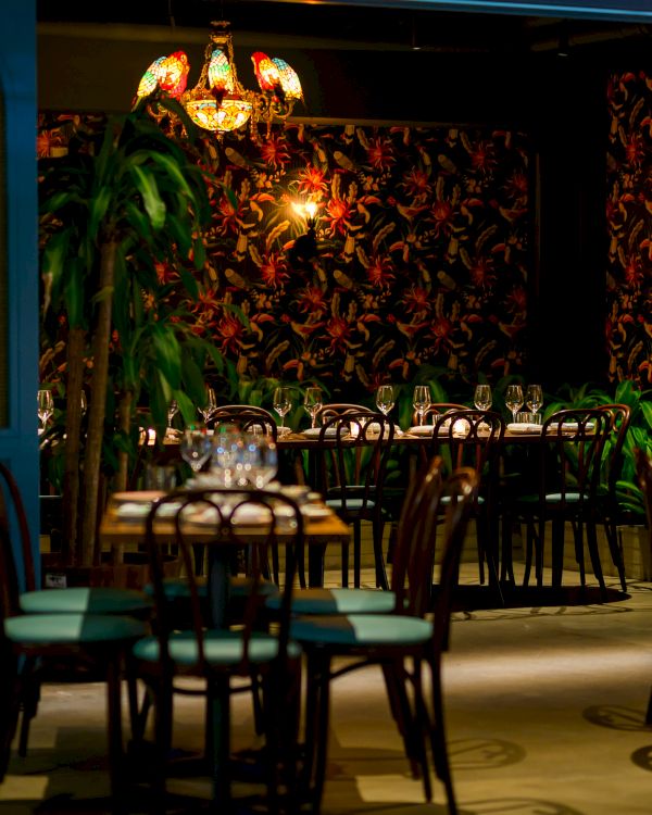 A stylish restaurant interior with dim lighting, patterned wallpaper, plants, and neatly set tables. Top it at 140 characters, always ending the sentence.
