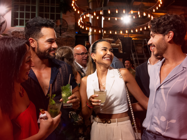 A group of stylish friends cheers with colorful cocktails at a lively indoor party under warm string lights, smiling and chatting in a festive vibe.