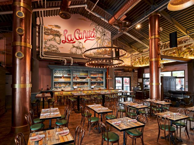 A stylish, industrial-style restaurant with copper columns, wooden tables and green chairs, a mural backdrop and a cozy bar area.