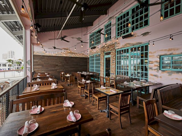 An empty rustic restaurant patio with wooden tables, pink napkins, string lights, teal window shutters, and ceiling fans&mdash;ready for guests.