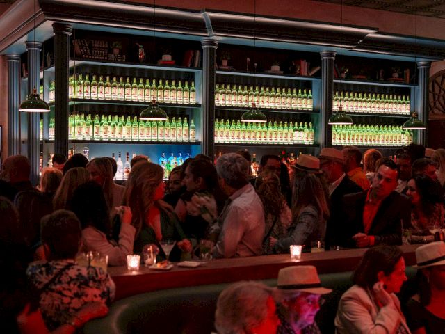 A crowded bar with shelves of illuminated bottles behind the counter, warm lighting, and people socializing at tables and the curved bar.