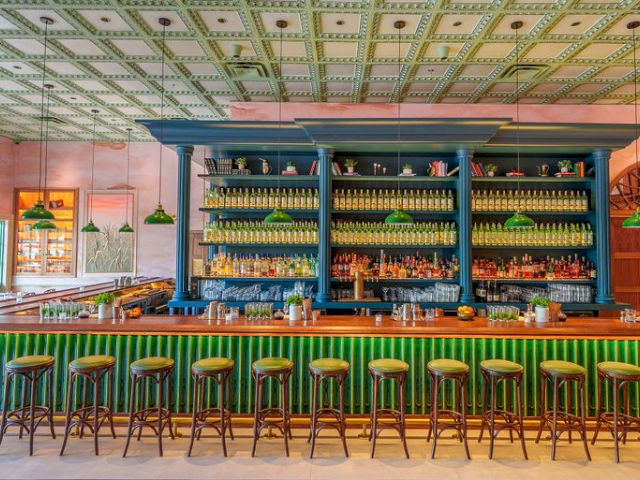 A stylish bar with a blue center shelf stocked with bottles, green stools along a long green counter, warm lighting, and arched windows in a colorful dining space.